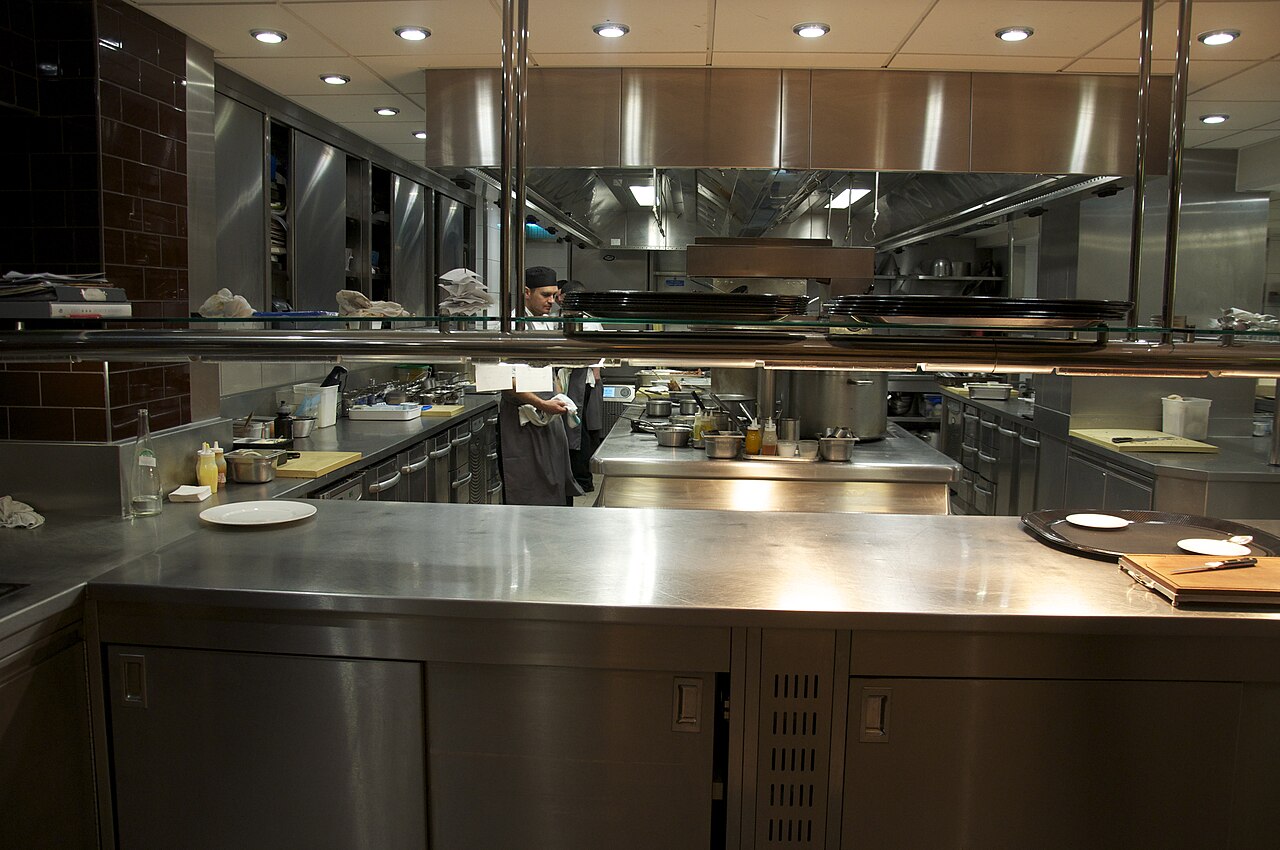 Professional commercial restaurant kitchen with stainless-steel equipment and large overhead extraction hoods.